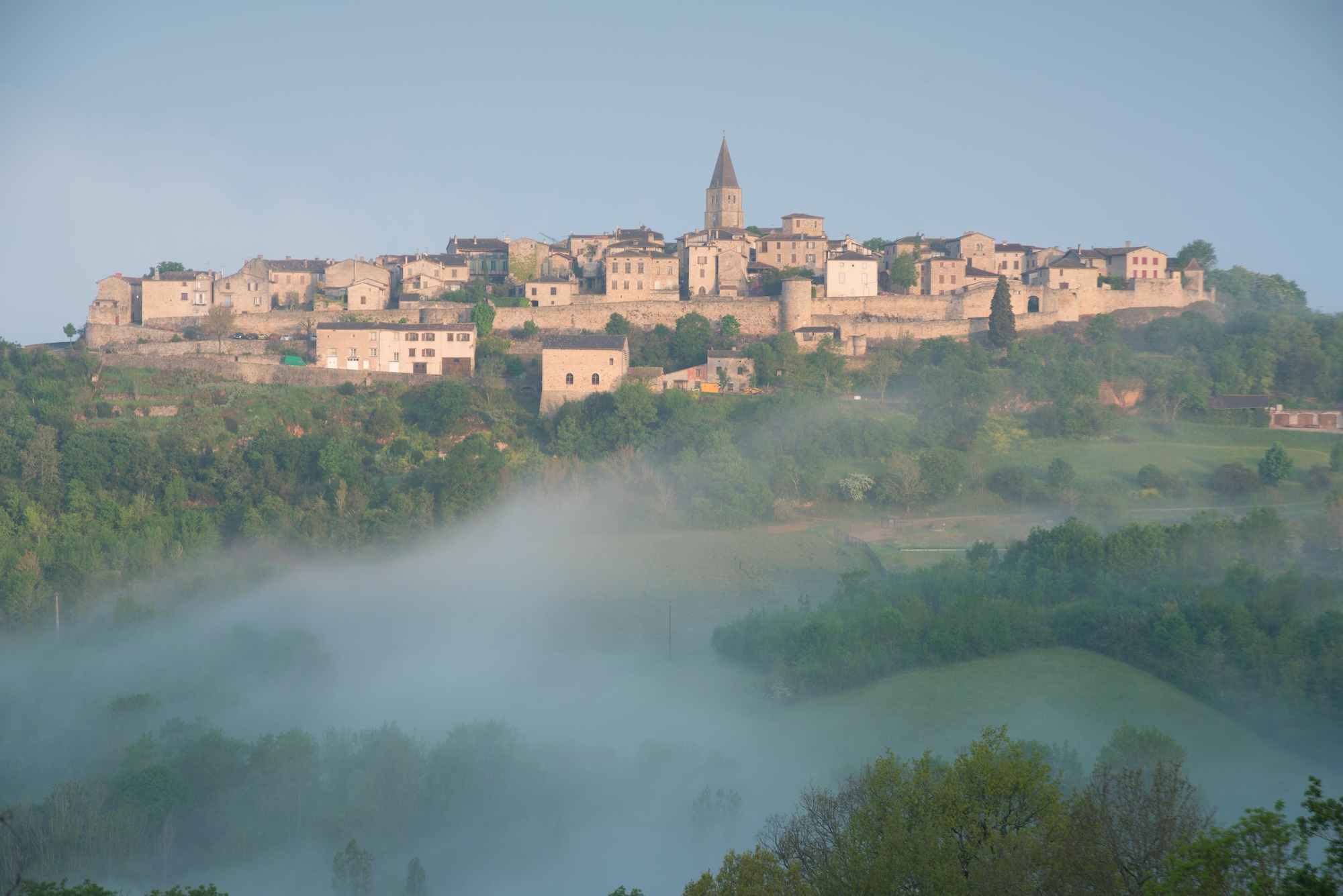 High-angle landscape view of an elegant European village nestled in a green mountain valley with lush forests and hills under a blue sky